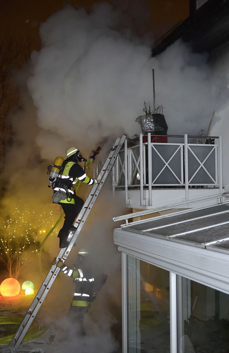 feuerwehrmann auf leiter mit atemschutzmaske vor balkon einer brennenden Wohnung mit viel Qualm und Rauch — © Berufsfeuerwehr München