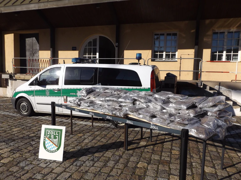 Sichergestelltes Marihuana auf einem Tisch. Im Hintergrund ein Zoll-Fahrzeug — © Foto: Zoll München