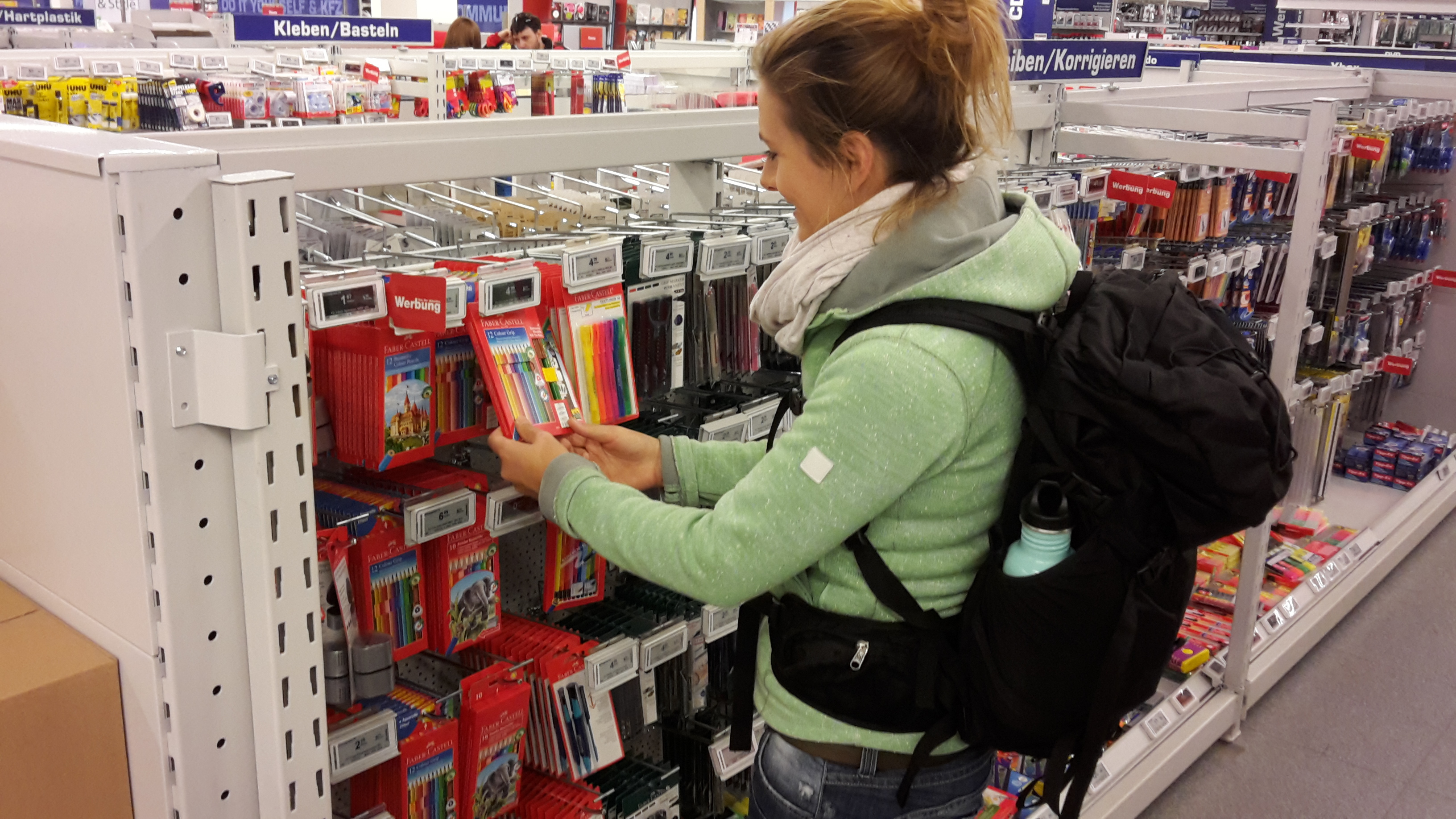 lena starkl im geschäft vor einer großen außwahl buntstifte und hält eine stifte packungin der hand, Nachrichtenvolontärin Lena Starkl beim Stiftekaufen